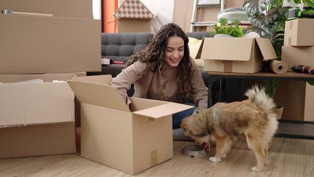 Young hispanic woman with dog unpacking cardboard box sitting on floor at new home