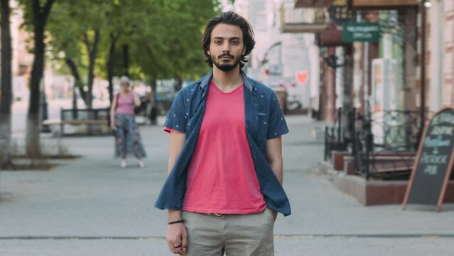 Time lapse portrait of handsome Arab man in casual clothing standing outdoors in city street alone with serious face and looking at camera while people are moving around.