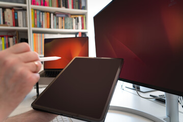 Tablet screen view. Hand holding a graphic pen. Computer and monitor display screens in background. Screens mockup. Home office studio.