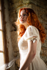 Fototapeta na wymiar Red-haired beautiful girl near the old window in a white night dress, in a French traditional house, the theme of romance
