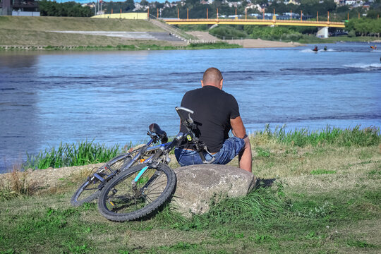 Man with bicycle sitting on o big stone looking at a river blue water and bike lying on grass near by