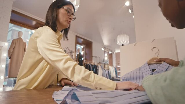 Young Caucasian woman bringing light blue striped shirt on hanger to Black female shop clerk at counter in designer showroom, examining its quality before purchase