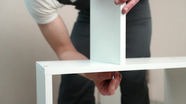 Furniture assembly. The furniture assembler takes a white shelf, tightens the screw with a hex wrench.