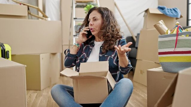 Middle age hispanic woman speaking on the phone unpacking cardboard box at new home