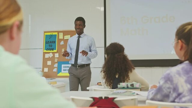 Enthusiastic young Black male teacher giving interactive IT lesson to group of diverse 6th grade students sitting at their desks in bright spacious classroom and learning to code