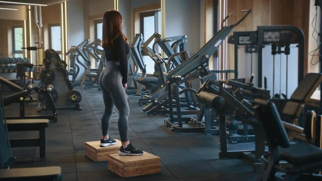 Athletic woman performing squats with kettlebell standing on wood blocks, feet shoulder-width apart. Gym equipped with strength training equipment. High quality 4k footage