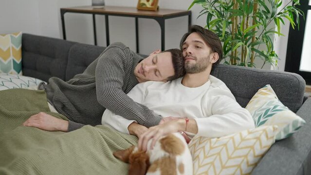 Two men couple hugging each other relaxing on sofa with dog at home
