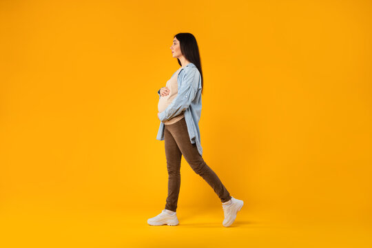Maternity concept. Pregnant lady touching belly and walking on yellow background, full length, copy space, side view