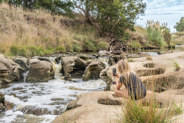 Woman taking photos with a mobile phone to a waterfall stream in nature.