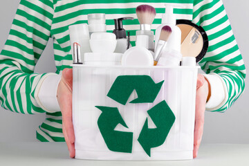A faceless woman is holding a container full of cosmetic products with a recycling sign. Used...