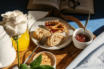 breakfast in bed, complete with coffee, pastries, orange juice, strawberry jam and flower. The breakfast tray in the warm glow of sunlight, accompanied by a book and eyeglasses on blue linens
