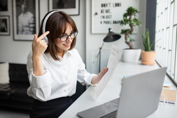 Cheerful young businesswoman having online meeting via laptop, holding papers and talking at computer camera