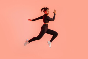 Fototapeta na wymiar Young sportive black woman training on red neon studio background, running and looking at camera, full length