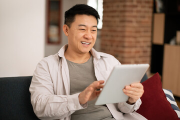 Relaxed happy asian mature man in casual outfit chilling on sofa at home, using digital tablet and smiling, copy space