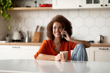 Cheerful Black Lady Drinking Coffee And Talking On Cellphone In Kitchen