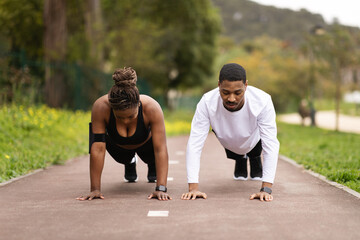 Obraz premium Serious young black couple do push-ups and look at phone, enjoy workout in summer park