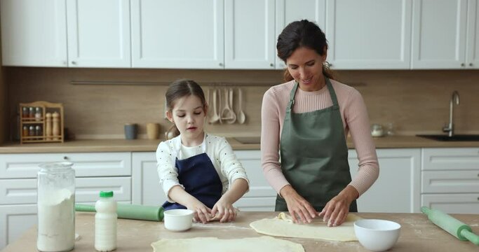 Young loving mother teach little 6s daughter to prepare dough, cooking together breakfast in kitchen. Pretty girl and mom wear aprons make home-made patries for family dinner. Share knowledge, love