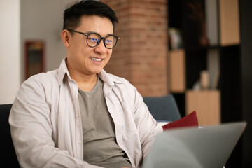Cheerful asian middle aged man in glasses with laptop working online, watch video lesson, blog,...