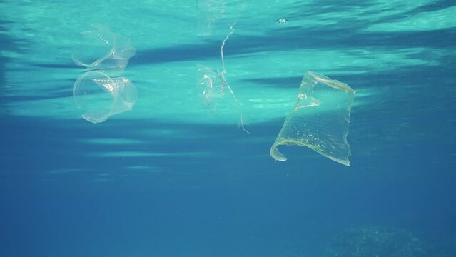 Close-up of plastic debris drifts under surface in blue water, Slow motion. Disposable plastic cups and pacs littering oceans of planet. Ecological problems, defending environment from pollution. 