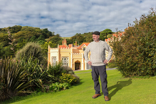 Man wearing a tweed cap and a sheep's wool jumper on a lawn in front of a typical southern England house. A sunny day with clouds in the sky.