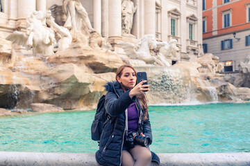 Content young Latin woman tourist in warm clothes with backpack and professional photo camera taking selfie on smartphone while sitting on border of Trevi Fountain during vacation in Rome, Italy