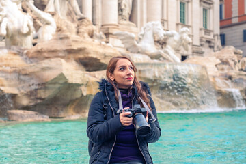 Content young Latin woman tourist in warm clothes sitting on border of Trevi Fountain with...