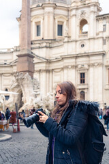 Trendy young Latin woman tourist in sunglasses and warm clothes standing with professional photo camera looking away in Piazza Navona in Rome, Italy