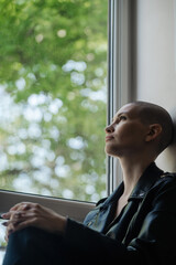 Beautiful young woman with a shaved head without hair sits and looks out the window
