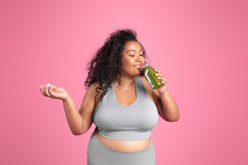 Positive black plus size woman in sportswear holding jar of fresh smoothie, standing on pink studio...