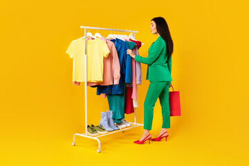 Stylish lady standing near garment rack and choosing new summer outfit, buying clothes in showroom,...