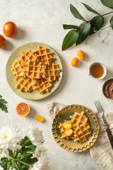 Plates with Belgian waffles with honey, chocolate sauce, bloody oranges and kumquat on a light background with white flowers. 