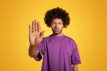 Serious confident mature black curly man in purple t-shirt making stop gesture with hand