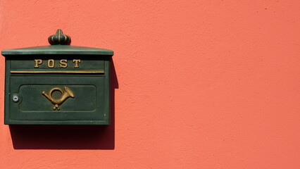 Green metal mailbox with golden post symbol on bright orange plastered wall in the sunshine. Space for text. 