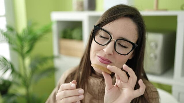 Beautiful hispanic woman eating cookie sitting on table at dinning room