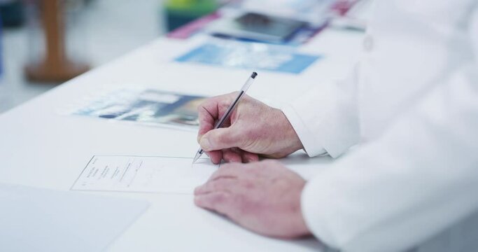 Man, doctor and hands writing prescription, diagnosis or medical certificate and note on desk at pharmacy. Hand of male healthcare professional filling in document, form or paperwork for application
