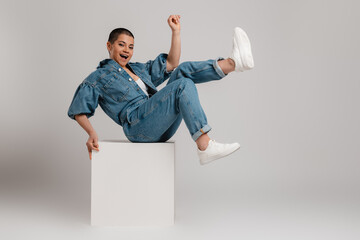 Playful young short hair woman in denim clothes having fun while sitting against grey background