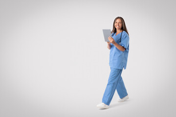 Friendly senior woman in medical uniform holding digital tablet, walking on grey background, full length, free space