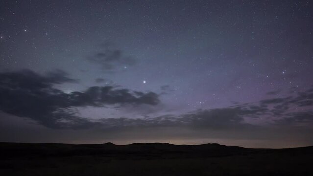 Nighttime lapse of a star filled sky with clouds moving from left to right and weak Aurora.  The distant horizon is a silhouette of rolling hills.