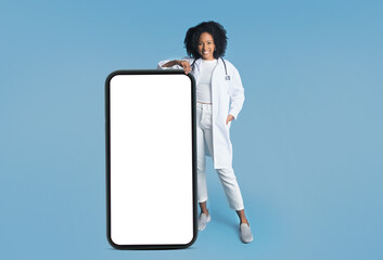 Glad millennial african american lady in white coat with stethoscope near big phone