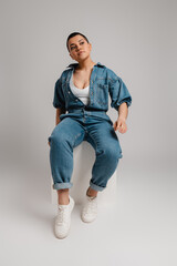 Fashionable young short hair woman in denim clothes sitting against grey background