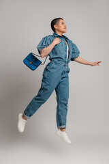 Young shaved head woman in denim clothes carrying bag while jumping against grey background