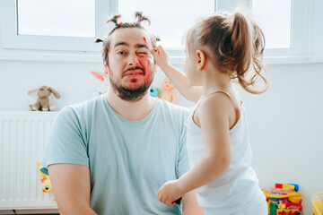 A father and daughter enjoying a funny and creative makeup session at home, with the dad...