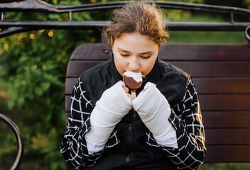 A girl, a child eats ice cream, holding in her hands with plaster, bandages after a bone fracture, feeling uncomfortable, sitting on a park bench. Photography, portrait.