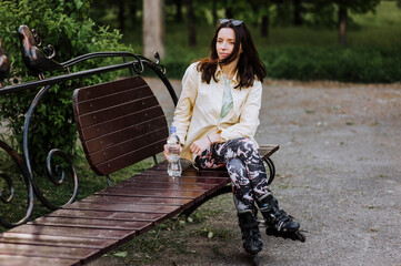 Beautiful young woman, girl athlete sits on a wooden bench in the spring park in roller skates with...