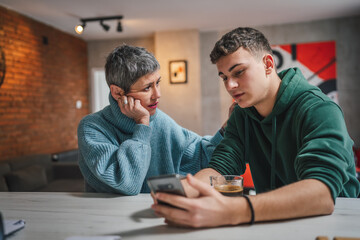 teenage boy and mature caucasian woman sit together at the kitchen at home talk mother and son or relatives support solving problem share experience and opinion boy hold mobile phone smartphone - Powered by Adobe