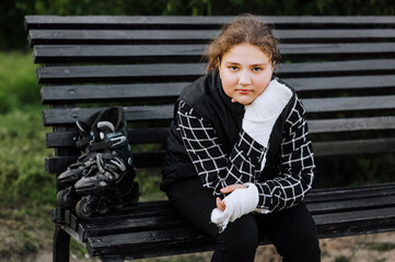 A girl in sadness, a child athlete with a plaster, a bandage on her hands after a bone fracture, sits on a bench in the park with roller skates, missing riding alone. Photography, portrait, sports.