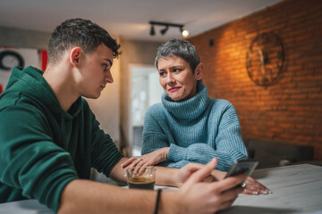 teenage boy and mature caucasian woman sit together at the kitchen at home talk mother and son or relatives support solving problem share experience and opinion boy hold mobile phone smartphone