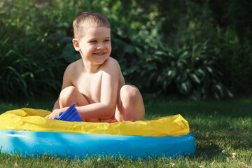 A smiling little boy plays in a yellow and blue rubber pool with a plastic toys,in background sunny backyard lawn and shade behind him