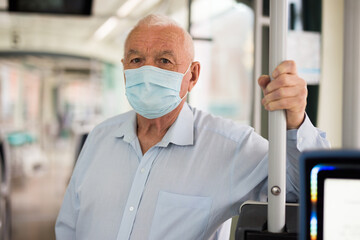 Elderly Caucasian man in mask standing in tram and looking in camera.