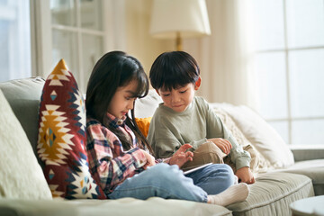 two cute little asian kids brother and sister sitting on family couch at home playing game using digital tablet